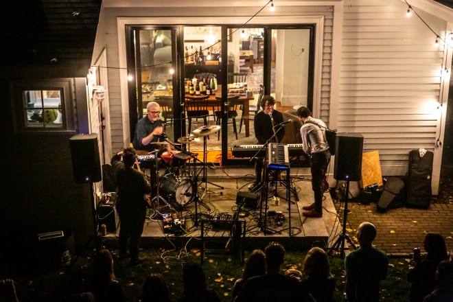 A small number of people gather below to watch a band play on a back porch.