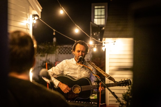 A musician sings passionately into a microphone while playing guitar, under hanging lights in a backyard. 
