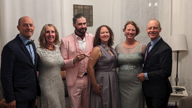 Three women and three men dressed in evening attire. The women are mostly wearing pastels, and one of the men is in a pepto bismol pink suit.