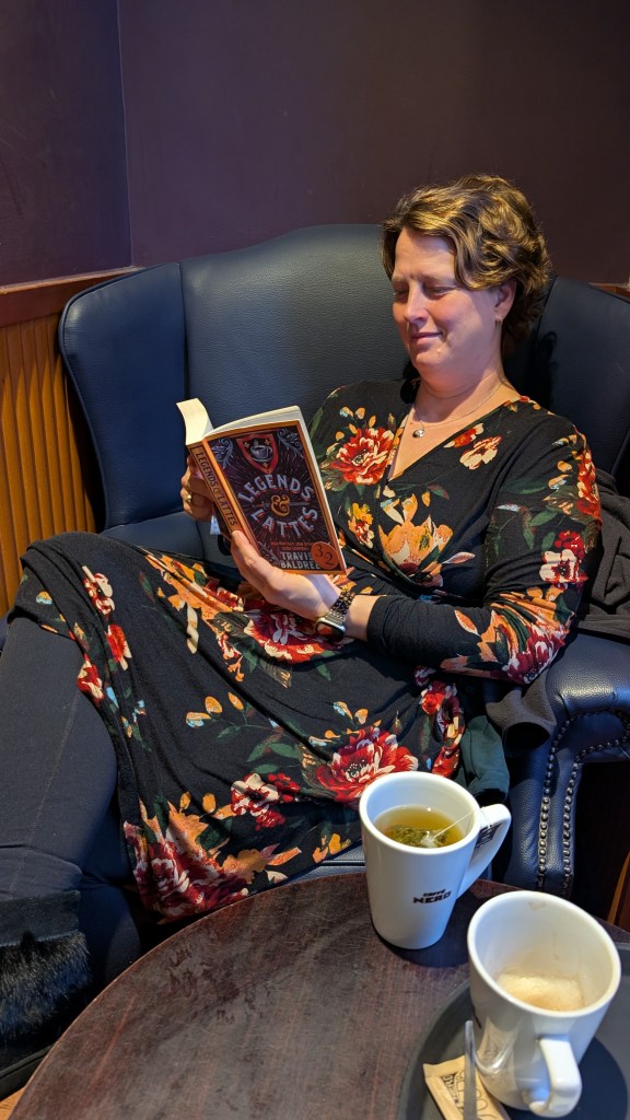 A middle aged woman in a floral dress happily reading in a blue armchair with a cup of tea in front of her.