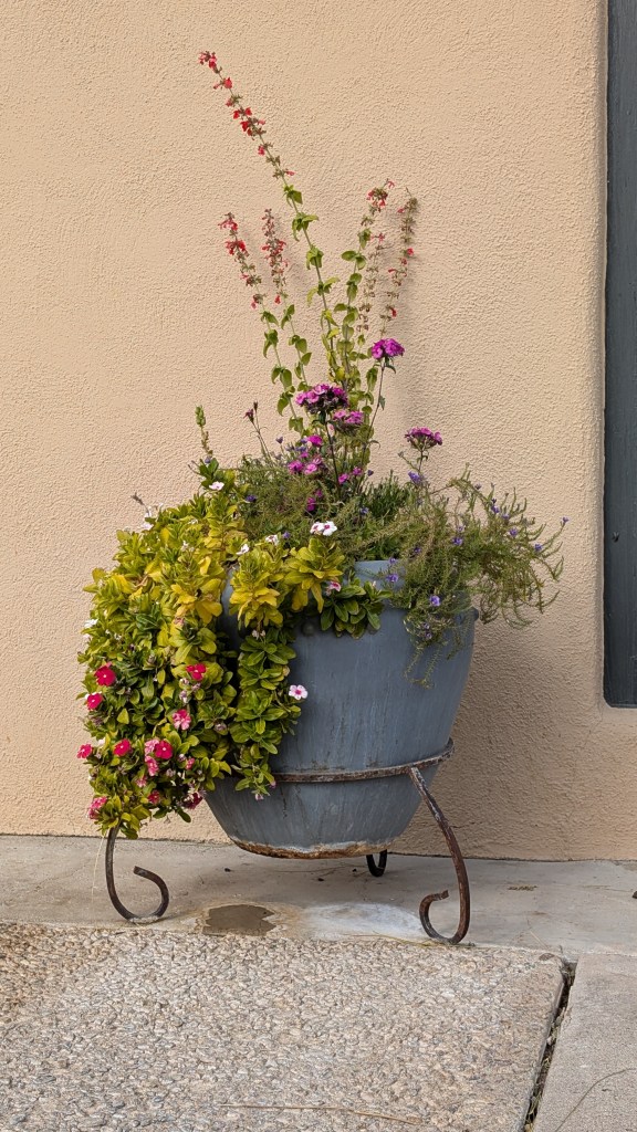 A beautiful pot on an iron stand full of greenery and flowers, against a warm beige wall