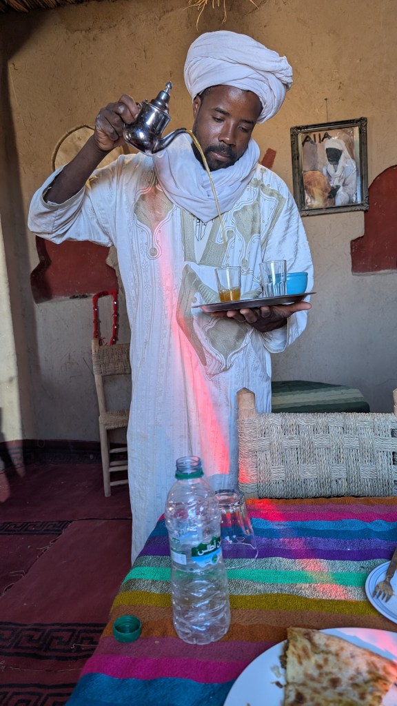 A black man in a white robe pouring mint tea from high up, with a strip of red light cutting across him like a sash