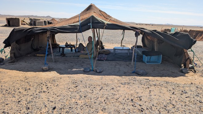 A low tent with only two walls, with sticks of wood and a roof of canvas. An American man sits in it and looks to the camera.