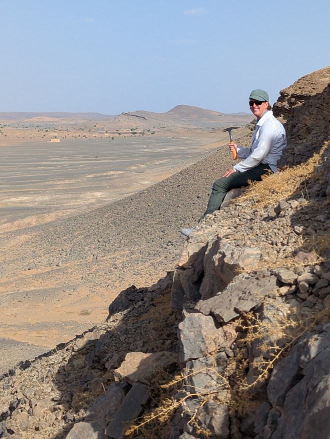 A woman holding a mining hammer on a very steep portion of a desert mountain