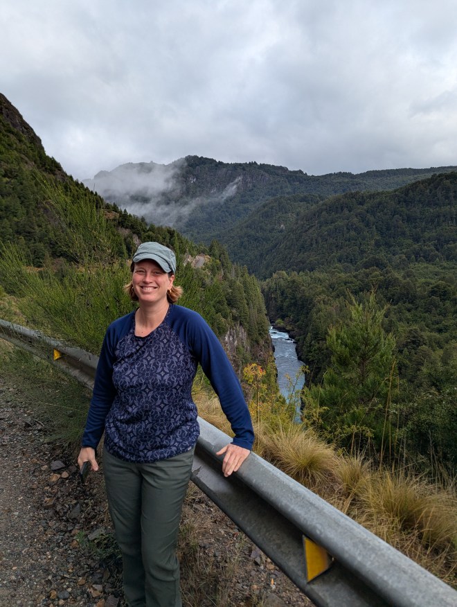 A woman standing with her hand on a guard rail. There are misty mountains behind her, and thin blue ribbon of river with whitewater cutting through a forest.