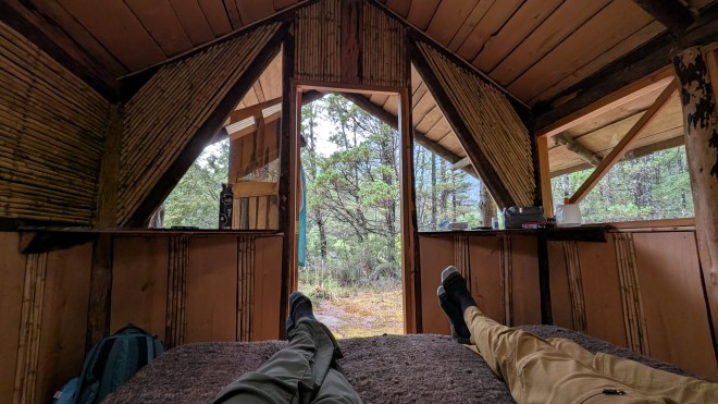 Two pairs of feet on a bed pointing at the open door of a small wooden cabin