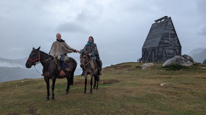 Two people wearing ponchos on horses - with a strange wooden building behind them. The scenery is wreathed in clouds.