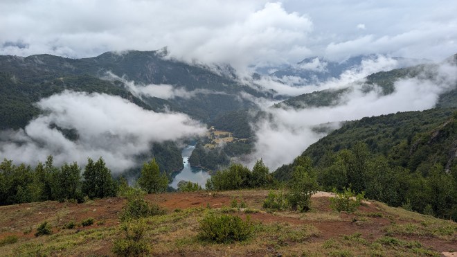 A high meadow looking far down at a blue river, with a rapid in it. You can see the clouds wreathing green mountains on every side.