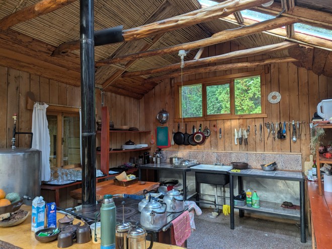 A rustic kitchen with a wood stove, sink and counters. There are knives and pans on the wall, and breakfast drinks on the counter.