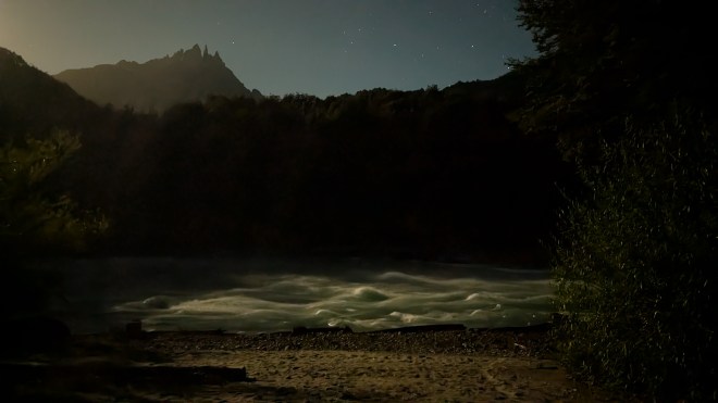 An eerie view of a mountain in dark sillouette with mooncaught whitewaters in the foreground and stars bright in the sky