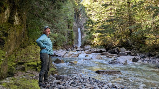 A woman standing on the banks of a stream with a dramatic waterfall behind