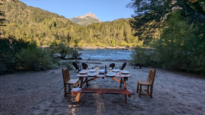 A table set in front of a fire, outside on the banks of a blue river, with spectacular mountains behind