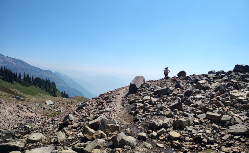 A small human figure silouetted against a blue sky, standing on a rocky terrain with distant mountains behind.