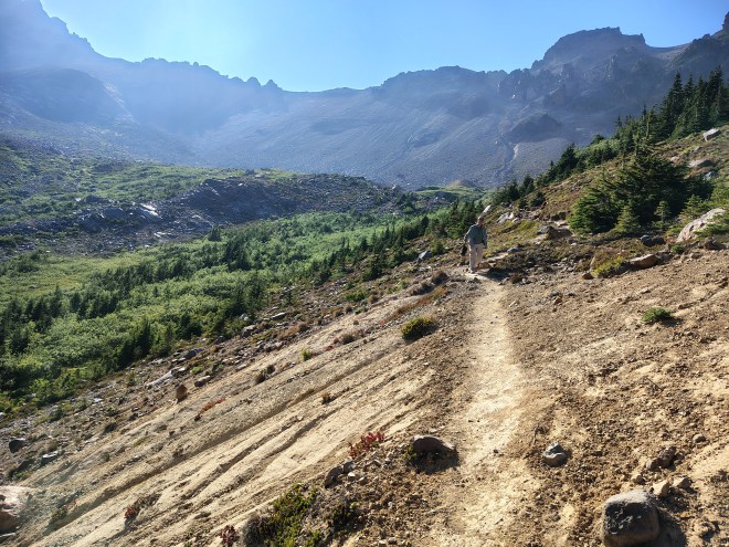 A very steep mountain path with a strong falloff to one side. A woman is walking carefully down. Huge mountains loom in the backgroun.