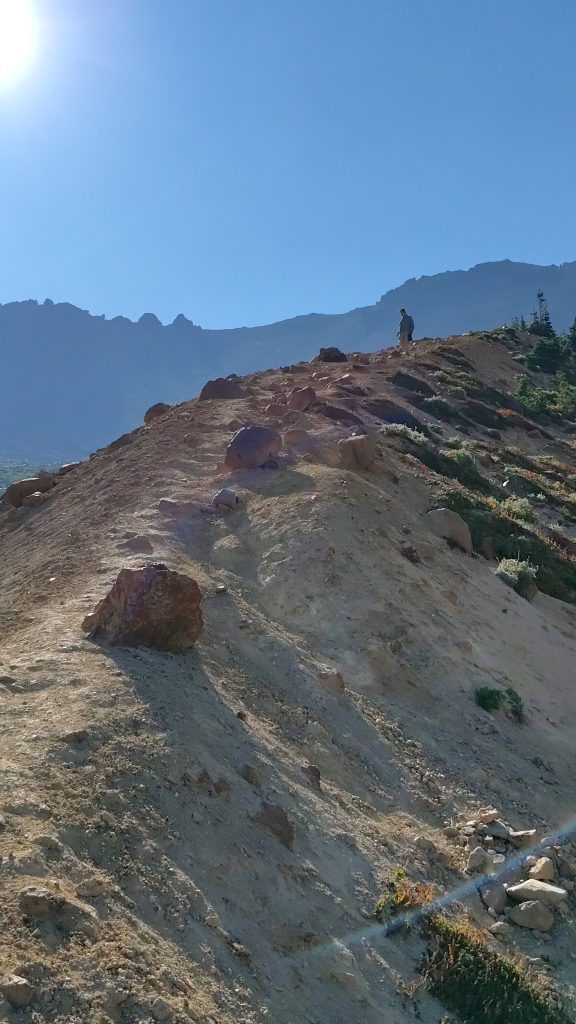 A woman standing at the top of a very steep trail that falls precipitously off to both sides. Glaciated mountains stand to either side